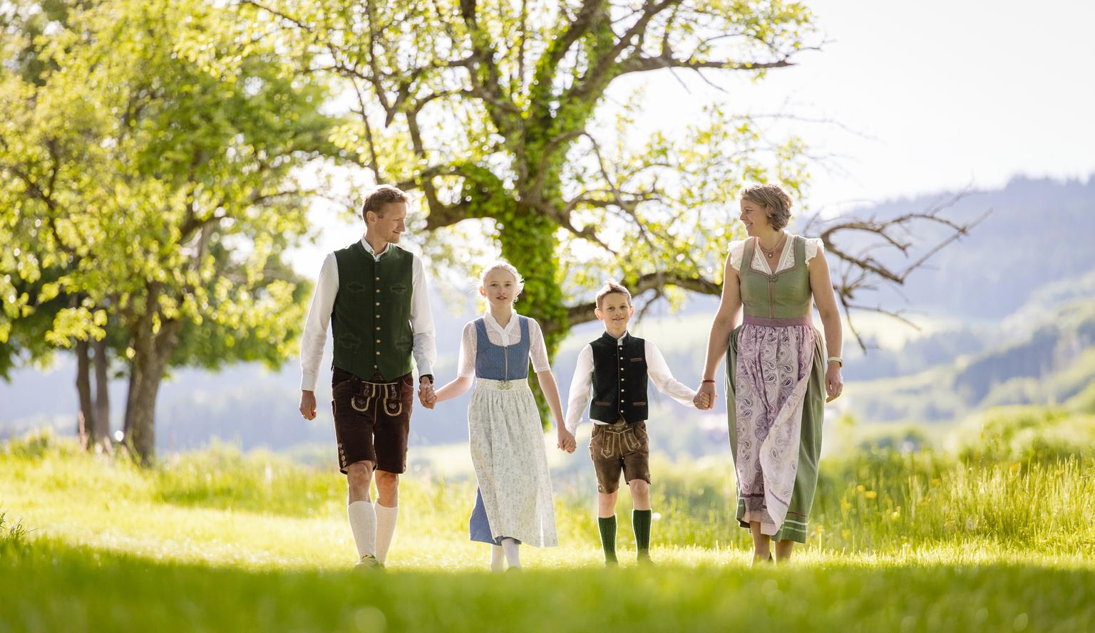 Eine Familie aus vier in Trachtenkleidung gehen händehaltend auf einer Wiese mit Bäumen im Hintergrund.