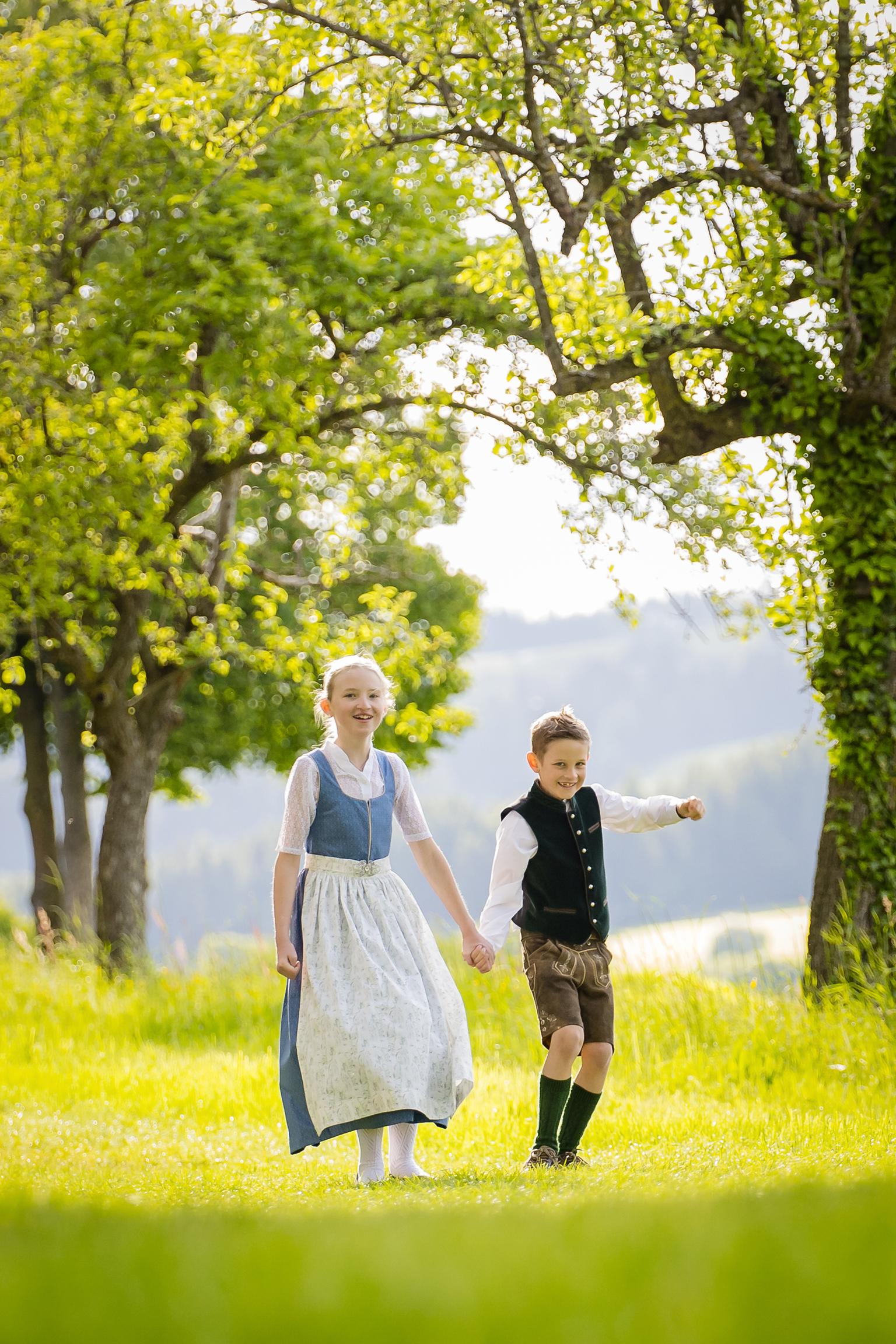 Zwei Kinder in Trachenkleidung gehen lächend auf einer Wiese mit Bäumen im Hintergrund.