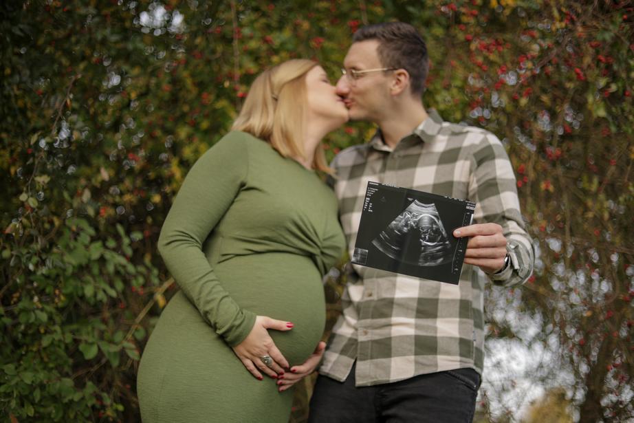 Eine schwangere Frau küsst einen Mann der ein Ultraschallbild in die Kamera hält.